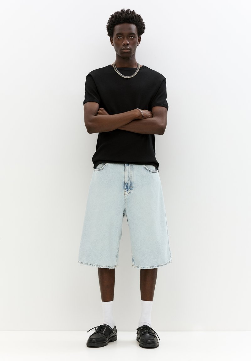 Tricou scurt negru cu mânecă scurtă, șorturi bermude din denim albastru deschis, pantofi negri și șosete albe. Modelul stă cu brațele încrucișate.
