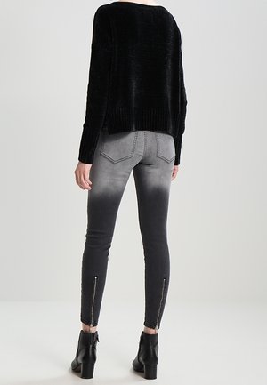 Person trägt graue bis schwarze eng anliegende Jeans mit Rückentaschen und Reißverschlüssen am Knöchel, schwarzen flauschigen Pullover und schwarze Stiefeletten und steht.