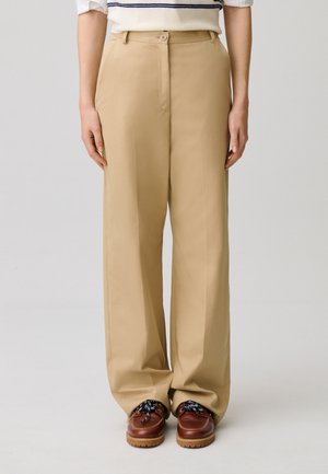 Pantaloni beige a gamba larga con bottone frontale e cerniera, indossati con scarpe marroni con lacci e un maglione bianco con righe blu navy.