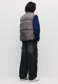 Grå puffer vest med høj krave, parret med en blå sweater og løse sorte cargo bukser. Hvide sneakers fuldender outfittet.