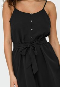 Combinaison sans manches noir avec un col rond, fermeture à boutons et une ceinture en tissu nouée à la taille. Matière douce et fluide avec une texture lisse.