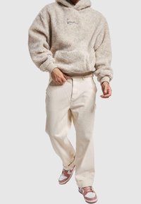 Uomo che indossa un hoodie beige in sherpa, pantaloni larghi color crema e sneakers bianche e rosa, in piedi contro uno sfondo semplice.