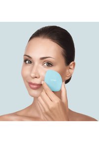 Applicatore per trucco in silicone blu chiaro, tenuto contro una pelle liscia, con una forma arrotondata. È visibile il nome del brand "GESKE".