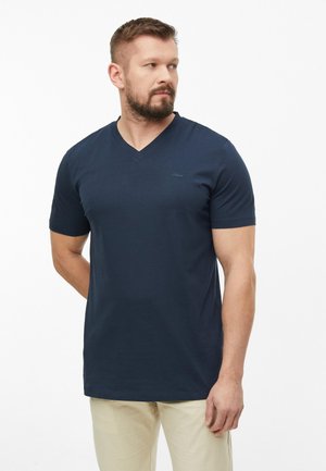 Mężczyzna z krótkimi włosami i brodą, ubrany w granatowy T-shirt z dekoltem w serek i jasnobeżowe spodnie, stoi na tle gładkiego białego tła.