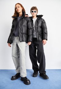 Două persoane îmbrăcate în jachete puffer negre stau în fața unui fundal deschis. Una poartă pantaloni gri, iar cealaltă pantaloni negri, ambele având încălțăminte masivă neagră.