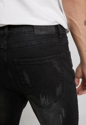 Jeans de mezclilla negra con corte ajustado, detalles desgastados y un parche de cuero en la cintura. Costuras expuestas y bolsillos traseros visibles.