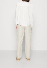 Blusa bianca con schiena arricciata, maniche lunghe, abbinata a pantaloni cargo beige chiaro con tasche laterali e vestibilità ampia. Sono indossati sandali col tacco.