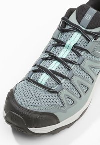 Les chaussures de trail présentent une tige en mesh gris clair avec des accents gris foncé, une semelle noire et des lacets turquoise. Embout renforcé et design texturé.