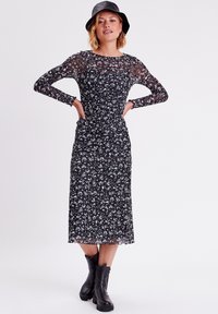 Robe noire à fleurs avec de longues manches transparentes, corps froncé et jupe mi-longue. Assortie à des bottes noires à hauteur de cheville et un chapeau noir.