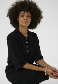 Vrouw met krullend haar in een zwarte geknoopte jurk, zittend met gekruiste handen, met een glanzende zilveren armband om haar rechterpols.