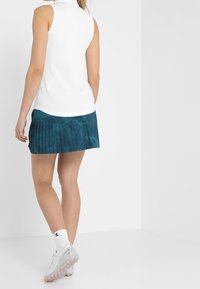 Bílý top bez rukávů s vysokým límcem spojený s tyrkysovou plisovanou sukní. Oblečení má hladkou texturu a sportovní designové prvky.