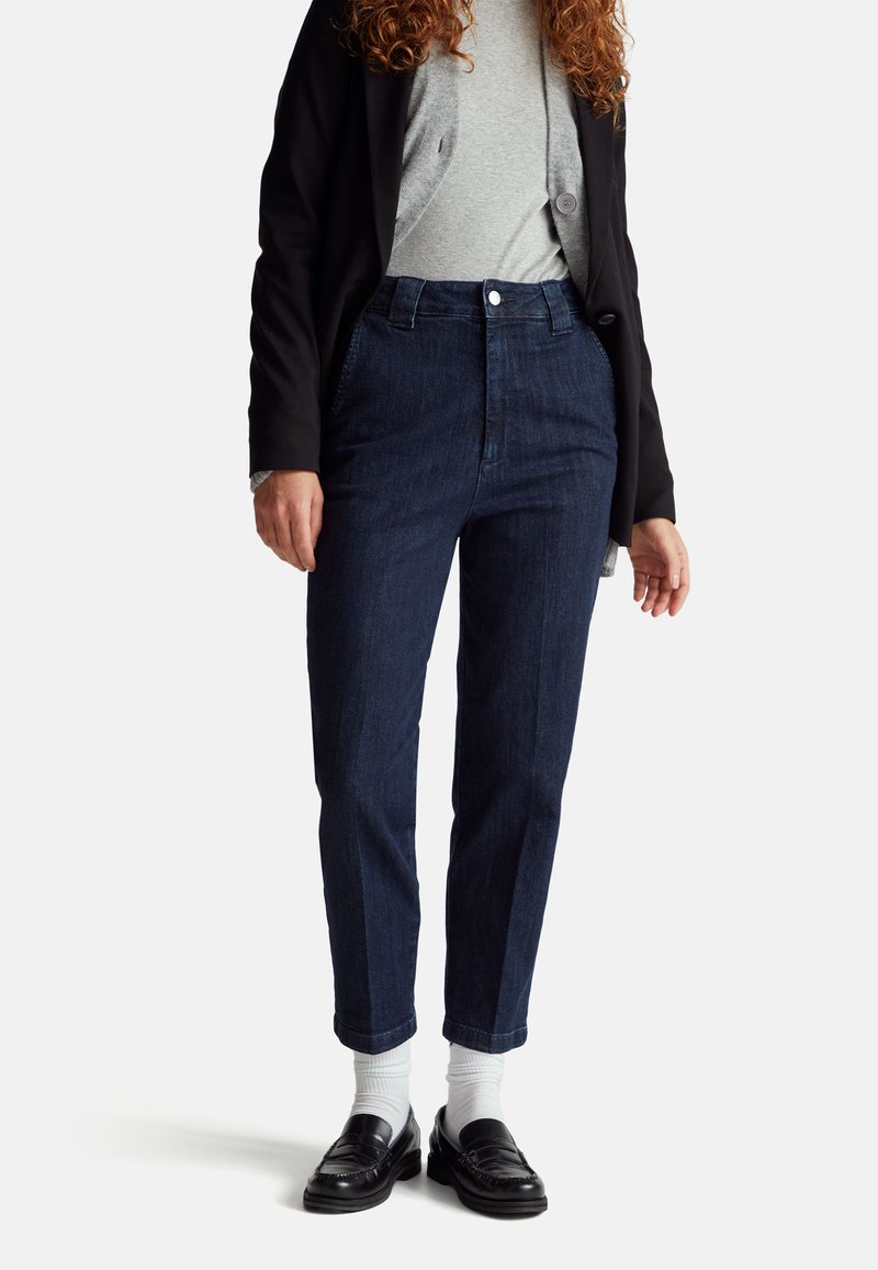 Ciemnoniebieskie spodnie z wysokim stanem o prostym kroju, w połączeniu z szarym topem i czarnym żakietem, uzupełnione czarnymi loafersami i białymi skarpetkami.