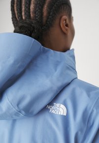 Personne aux cheveux tressés portant une veste bleu clair The North Face vue de dos, mettant l'accent sur la capuche et le logo.