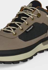 Μπεζ και μαύρο ορειβατικό παπούτσι Timberland με λευκά και κόκκινα κορδόνια με βούλες, μεταλλικά ρούχιστρα και παχιά σόλα με πέλμα.
