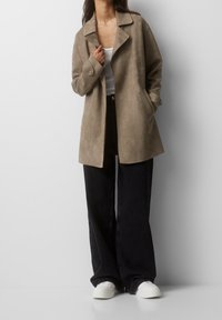 Mujer con chaqueta de ante beige, camiseta blanca sin mangas, vaqueros negros holgados y zapatillas blancas, de pie contra una pared blanca.