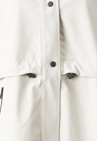 Lysebeige vandtæt jakke med høj krave, trykknapper og justerbar snor i taljen. Glat, mat tekstur og minimalistisk design.