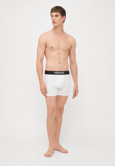 Fehér pamut boxer alsónadrág fekete gumírozott derékkal, amelyen a "VERSACE" logó található; testhez simuló design, sima textúra, comb középig érő hosszúság.