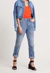 Veste en denim bleu clair, coupe courte ; chemise à col en v orange vif ; jean bleu clair taille haute avec revers ; sandales à talons noires.