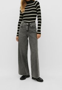 Jeans en denim gris à jambes larges avec une finition décolorée, associés à un haut à rayures noir et crème à manches longues en ribbed. Les chaussures sont des mocassins noirs.