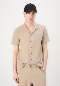 Camisa de manga corta beige claro con un acabado texturizado, cinco botones marrones y un bolsillo en el pecho. Se lleva con pantalones a juego y una gorra negra.