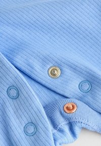Tessuto a coste di colore azzurro chiaro con bottoni a pressione: dettagli in oro, rame e blu. La texture è morbida con dettagli di coste visibili.