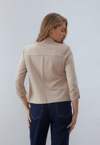 Vaalean beige takki, joka on viimeistelty muotoilu, pitkät hihat kääritty kyynärpäähän ja takasaumadetaili. Yhdistetty tummansinisiin farkkuihin.