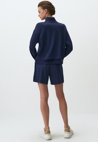 Completo sportivo blu navy composto da una maglia a maniche lunghe con colletto alto e dettagli in rete, abbinato a pantaloni shorts coordinati e scarpe da ginnastica bianche.