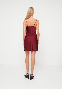 Bordo saten slip haljina s tankim trakama, dužine iznad koljena, glatke teksture i suptilnog A-linijskog oblika. Crne cipele s potpeticama s naglascima.