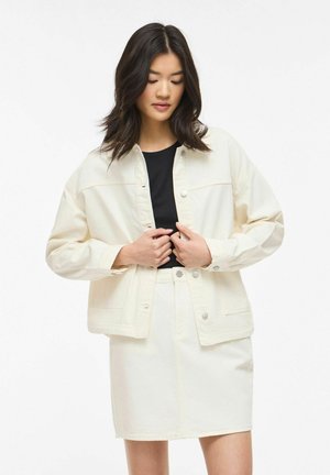 Noor nainen tumedate juustega, kes kannab helevalget jopet ja seelikut musta topi kohal, kinni nööpimas jopet mõlema käega.