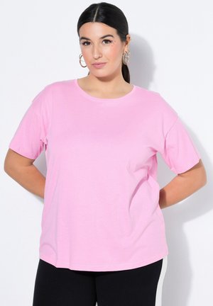 Frau mit dunklem Haar und Creolen, die ein rosa Kurzarmshirt und schwarze Hose vor weißem Hintergrund trägt.