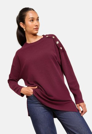 Burgundy ribbed sweater with an elongated hem and side slits, featuring gold buttons on one shoulder and long sleeves. Paired with blue jeans.