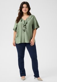 Blusa verde chiaro a maniche corte con scollatura a V profonda e bordi arricciati. Abbinata a pantaloni in denim blu e décolleté beige a punta.
