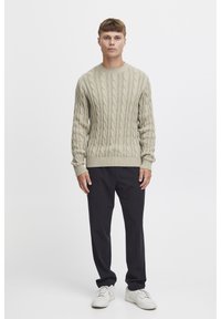 Pull en tricot beige avec des bords côtelés aux poignets et à l'ourlet, associé à un pantalon foncé et des baskets blanches, présentant une coupe décontractée et un motif texturé.