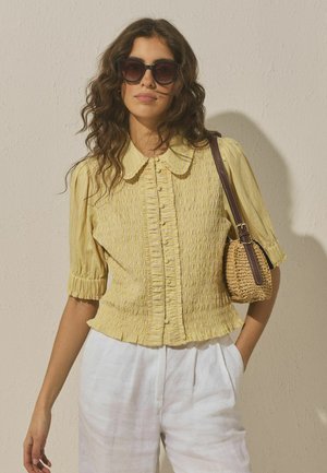 Sieviete ar viļņainiem matiem, valkājot saulesbrilles, dzeltenu teksturētu blūzi ar pogām, baltas bikses un turot austu somu pār plecu.