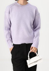Eine Person trägt einen lila gestrickten Pullover und schwarze Hosen, während sie mit einer Hand eine kleine weiße Handtasche an ihrem Griff hält.