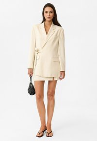 Crème-kleurige op maat gemaakte blazer met een tailleband, gecombineerd met een korte rok. Een zwarte mini-tas en sandalen met bandjes complementeren de outfit.