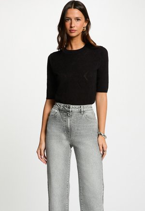 Femme aux cheveux bruns ondulés portant un haut noir à manches courtes avec un motif subtil, un jean taille haute clair délavé, des boucles d'oreilles créoles dorées et un bracelet argenté.