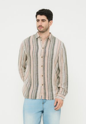 Mężczyzna ubrany w koszulę na guziki z długim rękawem w pionowe beżowe, szare i białe paski, w połączeniu z jasnoniebieskimi dżinsami, stojący na białym tle.