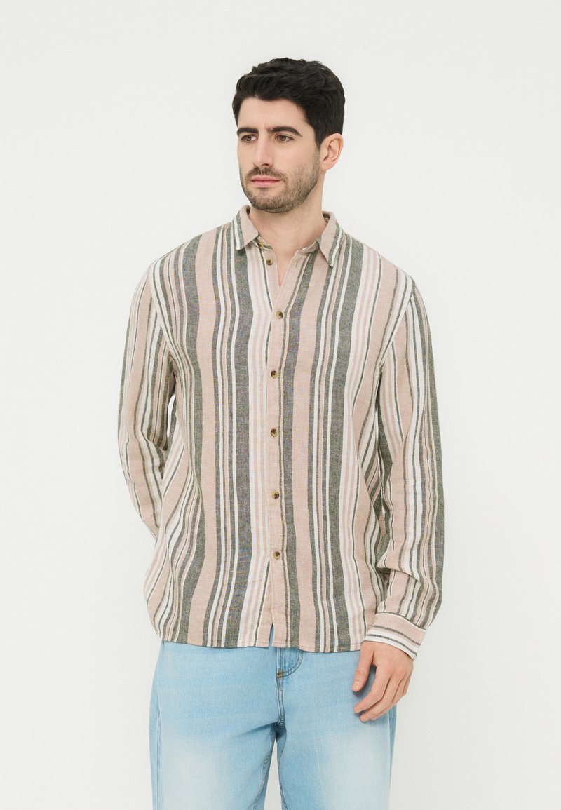 Άνδρας που φοράει πουκάμισο με μακριά μανίκια και κάθετες ρίγες σε μπεζ, γκρι και λευκό, συνδυασμένο με ανοιχτό μπλε τζιν παντελόνι, στέκεται μπροστά σε λευκό φόντο.