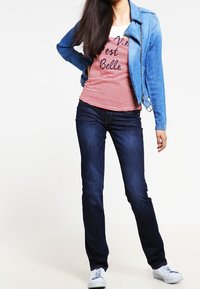 Giacca di denim blu chiaro, camicia a righe rossa e bianca con testo, jeans blu scuro, sneakers bianche. Modello in posa con una mano sui fianchi.