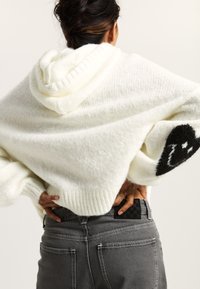 Un hoodie bianco oversize con orlo e maniche a costine. Design di un sorriso nero su una manica. Abbinato a jeans grigi con cuciture a contrasto.