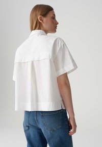 Weißes Kurzarmhemd aus leichtem Stoff, mit Rückenpasse und lässiger Silhouette, kombiniert mit dunkelblauen Jeans.