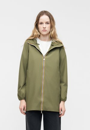 Ung kvinde med langt blondt hår iført en olivengrøn hættejakke med lynlås foran over en hvid skjorte og sorte bukser.