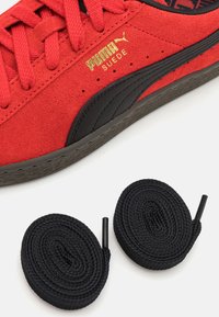 Crvene suede tenisice s crnim naglascima, s teksturiranom površinom, ravnim crvenim vezicama i crnim okruglim vezicama položenim sa strane. Vidljiv zlatni detalj s logom.