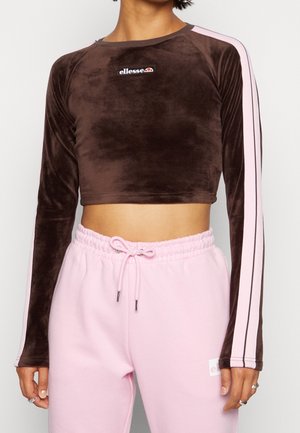 Braunes Samt-Crop-Top mit langen Ärmeln, das pinke Streifen entlang der Ärmel und ein kleines Logo auf der Brust aufweist. Kombiniert mit pinken Jogginghosen.