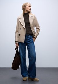 Beżowy płaszcz dwurzędowy na ciemnym swetrze z dzianiny, w połączeniu z niebieskimi spodniami o szerokich nogawkach i beżowym obuwiem, trzymając brązową torebkę z zamszu.