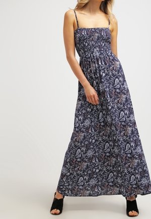 Frau trägt ein langes, dunkles Kleid mit floralem Muster, dünnen Schulterträgern und schwarzen offenen Schuhen, steht vor einem schlichten Hintergrund.