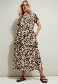 Maxi-mekko vaaleanbeige, jossa on musta aalto-kuvio, lyhyet hihat ja sivutaskut. Päällä mustat sandaalit, joissa on niittejä.