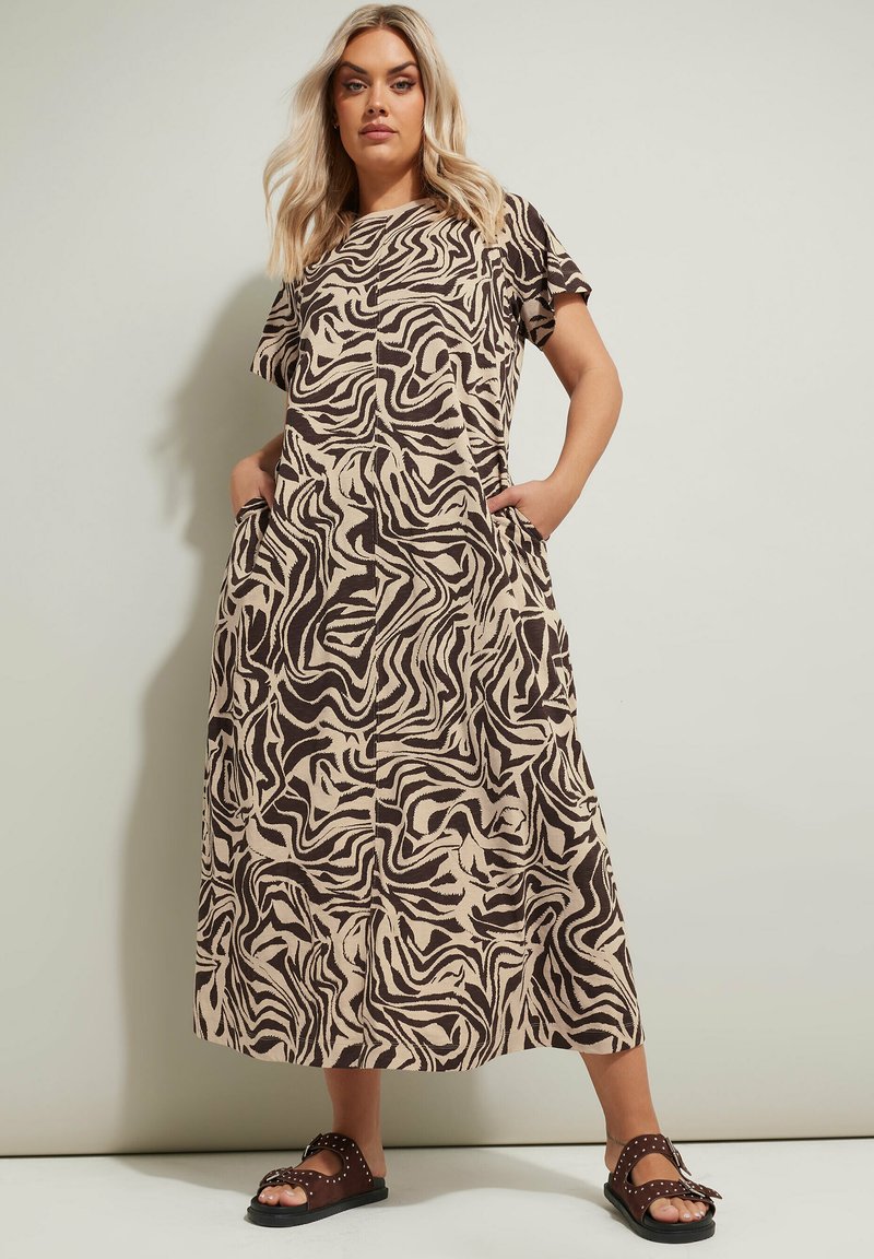 Maxi-mekko vaaleanbeige, jossa on musta aalto-kuvio, lyhyet hihat ja sivutaskut. Päällä mustat sandaalit, joissa on niittejä.