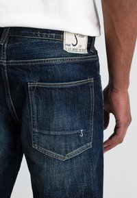 Donkerblauwe denim jeans met een nauwsluitend ontwerp, detailing op de achterzak en een label met de merknaam. Subtiele stikselaccenten.